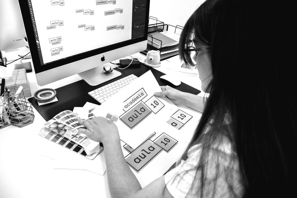 Foto en blanc i negre de la Laia treballant amb el logo de l'Acadèmia Aula 10 amb una carta de colors pantone i un iMac