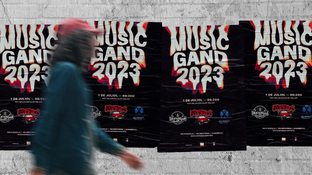 4 cartell del MusicGand 2023 enganxats a una paret de totxanes blanques i un senyor passant per davant