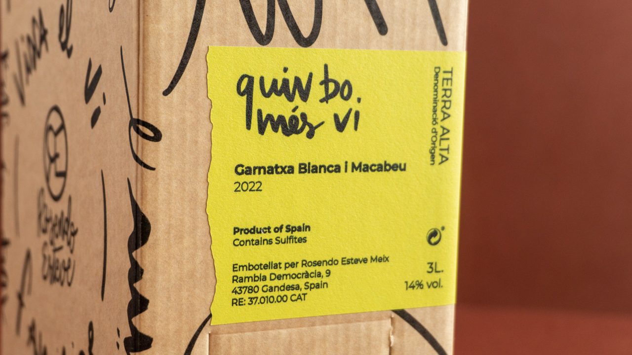 Detall de l'etiqueta de Quin Bo Més Vi en color groc, sobre la caixa en color kraft i un fons marró