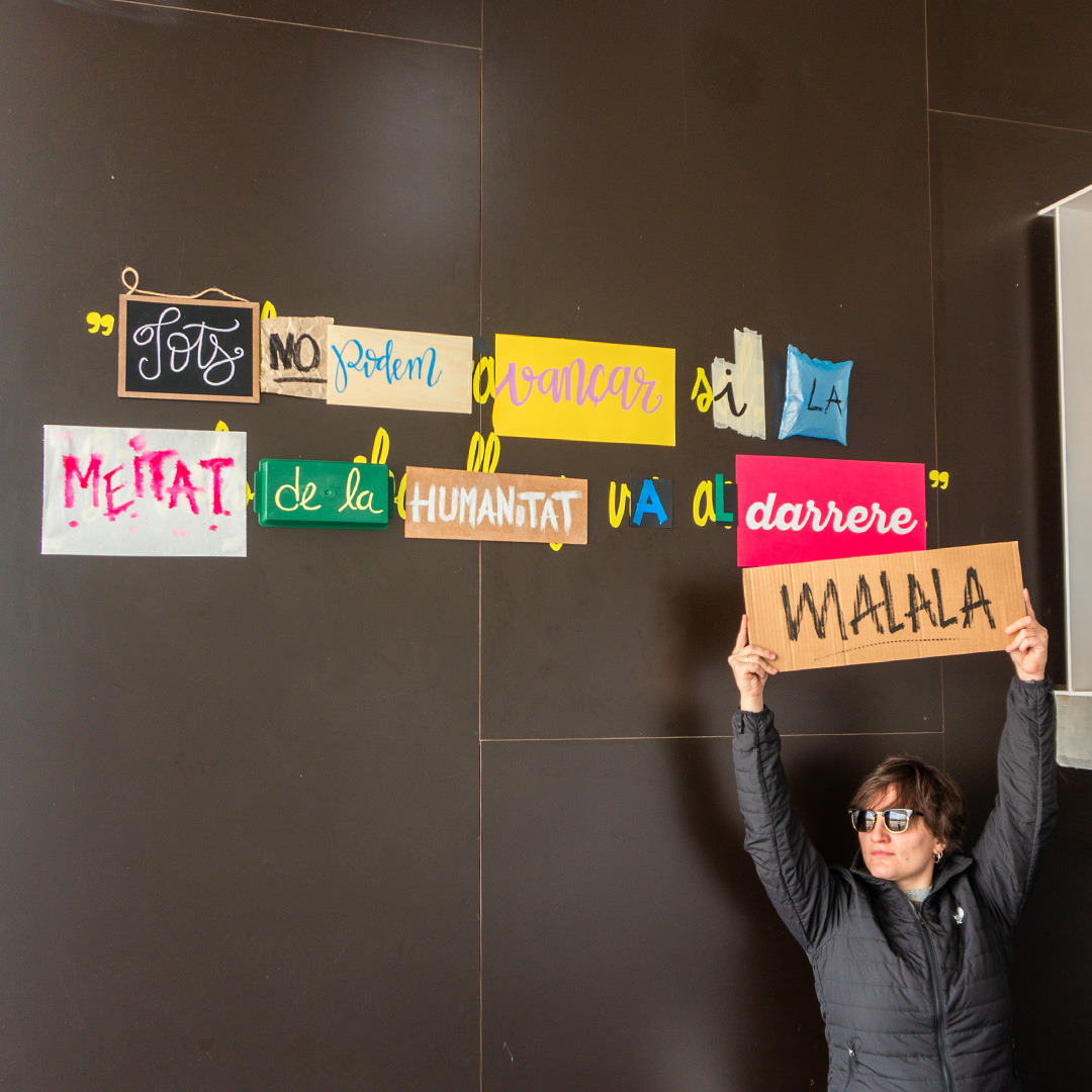 Frase de l'activista Malala creada amb diferents material i col·locada a sobre d'una paret marró i amb la Maite aguantant el cartell amb el seu nom